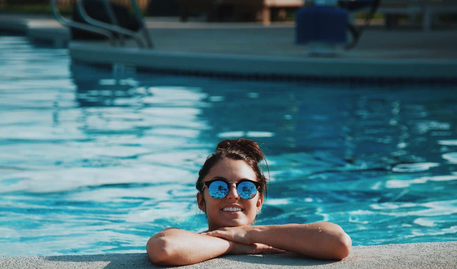 Sparkling swimming pool woman with sunglasses, in a pool, rests her arms on the ledge and smiles
